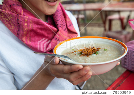 Cropped shot of woman holding a bowl with rice congee in her hand. Rice congee in Thailand known as 'chok' is a type of rice porridge served as breakfast topped with egg, pork, fried garlic etc. Cropped shot of woman holding a bowl with rice congee in her hand. Rice congee in Thailand known as 'chok' is a type of rice porridge served as breakfast topped with egg, pork, fried garlic etc. 79903949