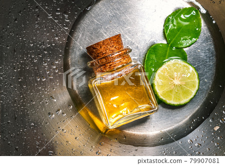 Packshot of Bergamot essential oil in a glass bottle with cork, fresh bergamot fruit, and green leaf on stainless steel with water drops background. Blank space on the bottle for label branding. 79907081