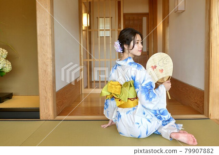 A young woman in a yukata sitting on a tatami mat and relaxing with a fan in her hand A young woman in a yukata sitting on a tatami mat and relaxing with a fan in her hand 79907882