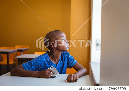 African american schoolboy holding face mask sitting at desk looking out of window in classroom African american schoolboy holding face mask sitting at desk looking out of window in classroom 79908709