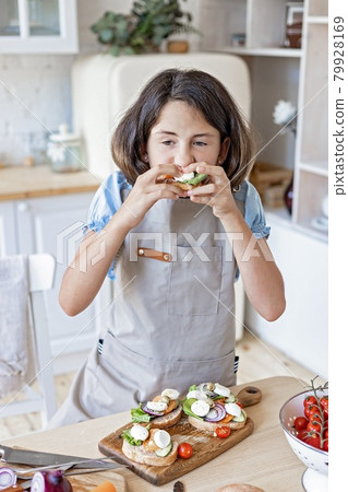 Cute caucasian teenage girl eating italian sandwiches at home kitchen. Beautiful girl prepares in the domestic kitchen. Attractive young girl in an apron on the background of the kitchen. Lifestyle Cute caucasian teenage girl eating italian sandwiches at home kitchen. Beautiful girl prepares in the domestic kitchen. Attractive young girl in an apron on the background of the kitchen. Lifestyle 79928169