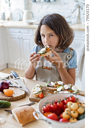 Cute caucasian teenage girl eating italian sandwiches at home kitchen. Beautiful girl prepares in the domestic kitchen. Attractive young girl in an apron on the background of the kitchen. Lifestyle Cute caucasian teenage girl eating italian sandwiches at home kitchen. Beautiful girl prepares in the domestic kitchen. Attractive young girl in an apron on the background of the kitchen. Lifestyle 79928170