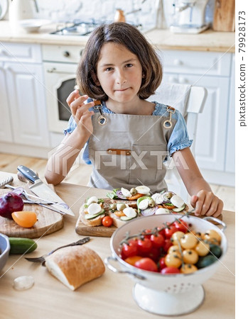Brunette teen makes sandwiches in the domestic kitchen. Beautiful girl is cooking in the home kitchen. Portrait attractive girl in apron on a light kitchen background. Looking at camera. Domestic life Brunette teen makes sandwiches in the domestic kitchen. Beautiful girl is cooking in the home kitchen. Portrait attractive girl in apron on a light kitchen background. Looking at camera. Domestic life 79928173