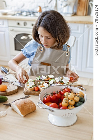 Portrait of a cute teenager cuts vegetables in the home kitchen. Beautiful brunette girl prepares in the domestic kitchen. Attractive young girl in an apron on the background of the kitchen. Lifestyle Portrait of a cute teenager cuts vegetables in the home kitchen. Beautiful brunette girl prepares in the domestic kitchen. Attractive young girl in an apron on the background of the kitchen. Lifestyle 79928174
