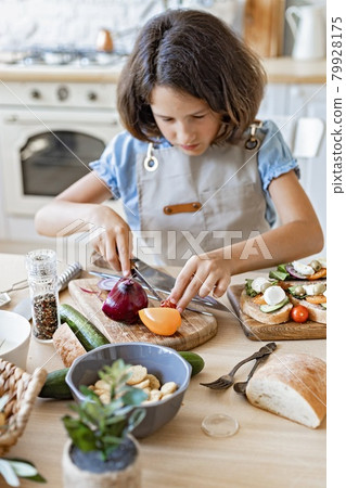 Portrait of a cute teenager cuts vegetables in the home kitchen. Beautiful brunette girl prepares in the domestic kitchen. Attractive young girl in an apron on the background of the kitchen. Lifestyle Portrait of a cute teenager cuts vegetables in the home kitchen. Beautiful brunette girl prepares in the domestic kitchen. Attractive young girl in an apron on the background of the kitchen. Lifestyle 79928175