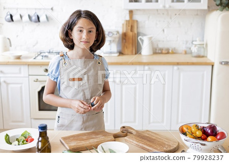 Beautiful cute teenage girl is cooking in the kitchen. Attractive girl in an apron on a light color kitchen background. Pretty brunette in the domestic kitchen. Copy space. Looking at camera. Beautiful cute teenage girl is cooking in the kitchen. Attractive girl in an apron on a light color kitchen background. Pretty brunette in the domestic kitchen. Copy space. Looking at camera. 79928237