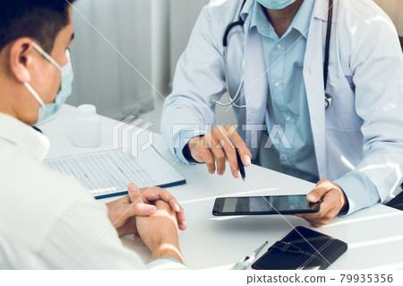 Doctors are explaining the treatment of a sick patient in tablets while wearing a mask during the virus outbreak. Doctors are explaining the treatment of a sick patient in tablets while wearing a mask during the virus outbreak. 79935356