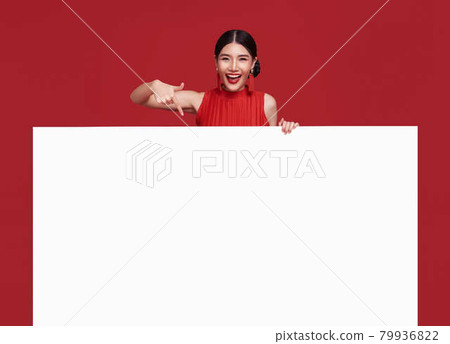 Happy asian woman in red casual attire with hand pointing blank announcement banner in red background. Happy asian woman in red casual attire with hand pointing blank announcement banner in red background. 79936822