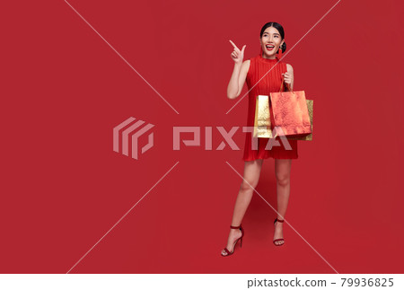 Happy Asian shopaholic woman wearing traditional cheongsam qipao dress hand pointing up to copy space isolated on red background. Happy Chinese new year 79936825