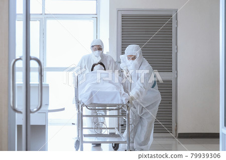 asian male nurse and doctor wearing ppe suit and face mask pushing stretcher gurney bed with seriously infected coronavirus or covid-19 patient towards quarantine room in hospital. 79939306