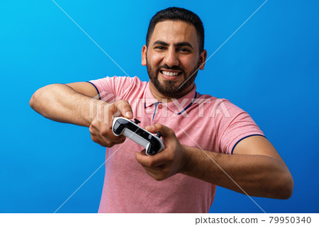 Young arab man in pink t-shirt playing video games against blue background Young arab man in pink t-shirt playing video games against blue background 79950340