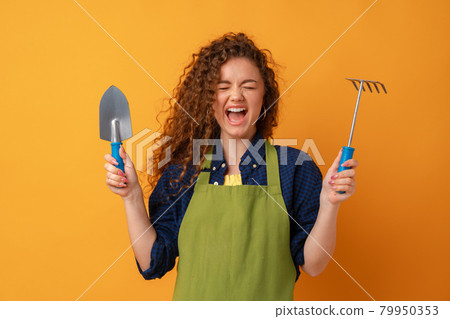 Young gardener woman wearing apron holding gardening tools against yellow background 79950353