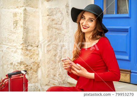 Portrait of tourist young caucasian woman in a red long dress and black hat with suitcase sitting on the stairs outdoors. Portrait of tourist young caucasian woman in a red long dress and black hat with suitcase sitting on the stairs outdoors. 79953270