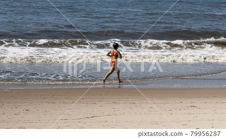 Female lifeguard in red bikini running in the ocean water on the edge of the beach 79956287