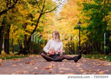 A young woman is sitting on the bench in the park and playing ukulele 79956944