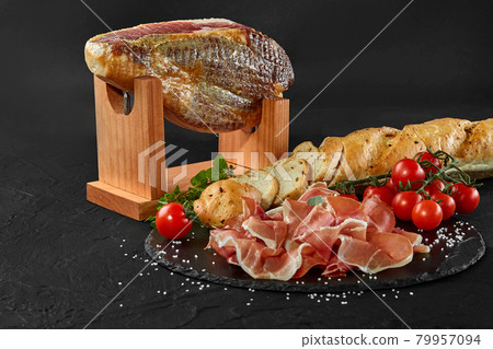 Jamon carved from pig leg on a jamonera stand, cherry tomatoes, baguette, herbs, spices on black slate board. Dark background. Close-up. Side view. Jamon carved from pig leg on a jamonera stand, cherry tomatoes, baguette, herbs, spices on black slate board. Dark background. Close-up. Side view. 79957094