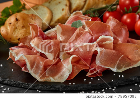 Sliced jamon, cherry tomatoes, baguette, herbs and spices on black stone slate board against a dark grey background. Close-up shot. Side view. 79957095