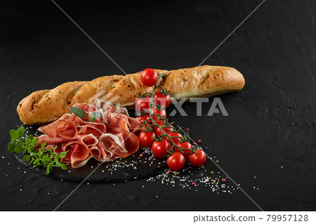 Sliced jamon, cherry tomatoes, herbs and spices on black stone slate board. Baguette is laying nearby on dark grey background. Close-up. Side view. Sliced jamon, cherry tomatoes, herbs and spices on black stone slate board. Baguette is laying nearby on dark grey background. Close-up. Side view. 79957128