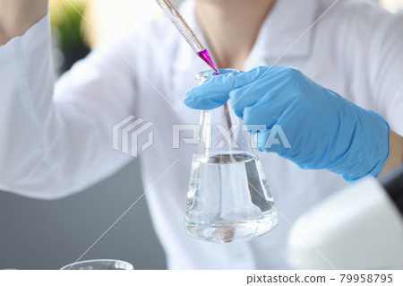 Gloved scientist holds flask of water and drops purple liquid from pipette into it 79958795