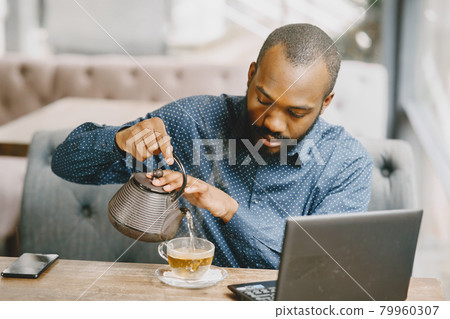 Man sitting in a cafe and working behind a laptop and pouring a tea in a cup. 79960307
