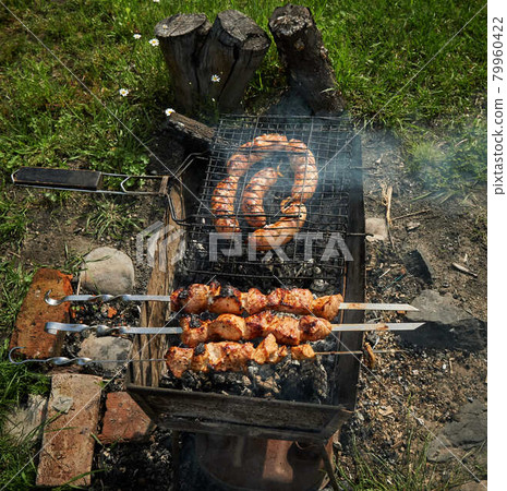 Barbeque meat and sausages or bratwurst on a grill grate in backyard. Man preparing shashlik or shish kebab over charcoal. Grilled meat on metal skewer outdoor. BBQ party or picnic food. Close up shot 79960422