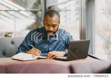 Man sitting in a cafe and working behind a laptop, writing in a notebook. 79960423