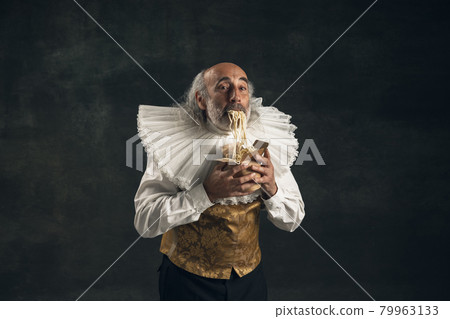 One elderly gray-haired man, medieval hystorical person, actor tasting fast food isolated on dark vintage background. Retro style, comparison of eras concept. One elderly gray-haired man, medieval hystorical person, actor tasting fast food isolated on dark vintage background. Retro style, comparison of eras concept. 79963133