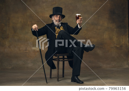 Elderly gray-haired man, gentleman, aristocrat or actor drinking beer isolated on dark vintage background. Retro style, comparison of eras concept. Elderly gray-haired man, gentleman, aristocrat or actor drinking beer isolated on dark vintage background. Retro style, comparison of eras concept. 79963156