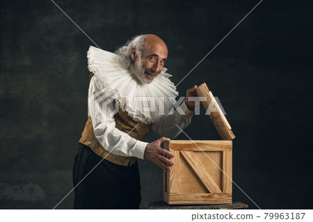 Elderly gray-haired man, actor or magician isolated on dark vintage background. Retro style, comparison of eras concept. Elderly gray-haired man, actor or magician isolated on dark vintage background. Retro style, comparison of eras concept. 79963187