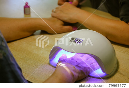 Woman hand inside UV LED lamp for drying pink nails with gel method, plate curing close up. Manicure process. Curing recently applied gel nail polish or hybrid varnish in beauty salon, beautician.  79965577
