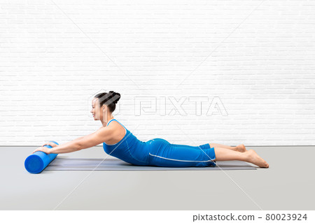 Pilates workout indoor. Caucasian attractive woman in blue sportswear lying on front and practice swan drill on a foam roller in studio, side view. 80023924