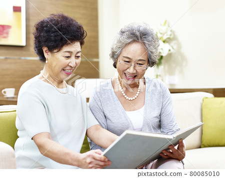 two senior asian women sitting on couch reading a book together 80085010