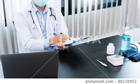 Medical concept a male doctor wearing a mask writing prescription to patient at his modern clinic for recovering from Coronavirus (COVID-19) 80110452