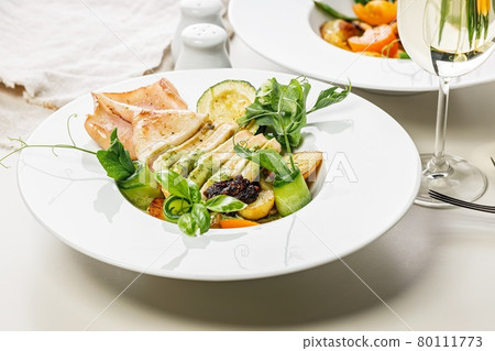 Close-up grilled squid with vegetables and a glass of white wine. Squid fillet baked with grilled vegetables with spicy green sauce. Delicious and healthy seafood. Mediterranean cuisine recipe 80111773