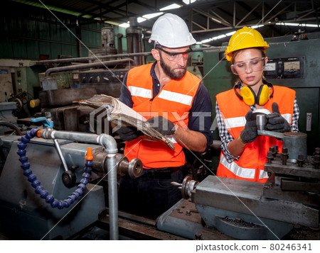 Confident young female mechanical engineer in a protective uniform, helmet while listening to colleague explanation workflow the CNC Machinery while working at the plant. Confident young female mechanical engineer in a protective uniform, helmet while listening to colleague explanation workflow the CNC Machinery while working at the plant. 80246341