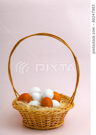 A rustic wicker basket with natural white and brown chicken eggs stands on a pink background. A rustic wicker basket with natural white and brown chicken eggs stands on a pink background. 80247983