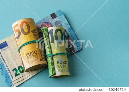 Roll of 100 euro banknotes and roll of 50 euro bills over 20 euro paper against blue background.  80249714