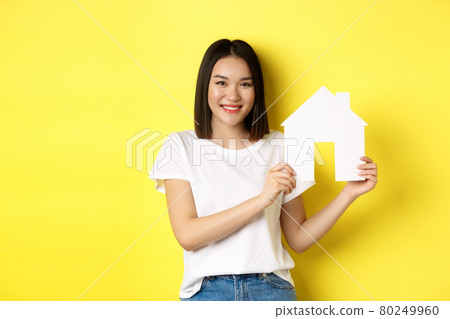 Real estate concept. Smiling beautiful woman showing paper house cutout and looking at camera, buying property, standing over yellow background Real estate concept. Smiling beautiful woman showing paper house cutout and looking at camera, buying property, standing over yellow background 80249960