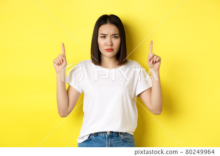 Beauty and fashion concept. Beautiful asian woman in white t-shirt pointing fingers up, standing over yellow background Beauty and fashion concept. Beautiful asian woman in white t-shirt pointing fingers up, standing over yellow background 80249964