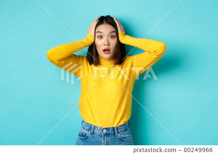 Shocked asian woman looking speechless, gasping amazed and holding hands on head, hear big news, standing over blue background 80249966