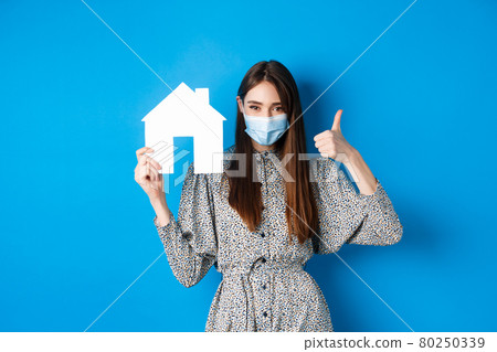 Real estate, covid-19 and pandemic concept. Candid woman in medical mask showing thumb up and paper house cutout, reanting apartment, standing on blue background Real estate, covid-19 and pandemic concept. Candid woman in medical mask showing thumb up and paper house cutout, reanting apartment, standing on blue background 80250339