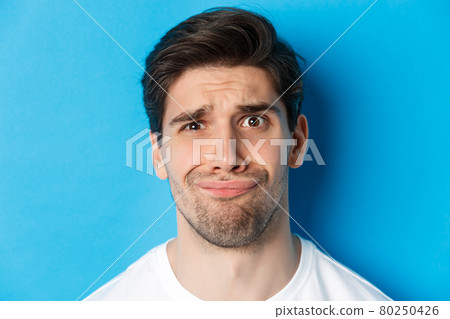 Head shot of skeptical and disappointed man, grimacing complicated, standing against blue background 80250426