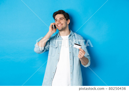 Smiling man call bank support, talking on mobile phone and holding credit card, standing against blue background Smiling man call bank support, talking on mobile phone and holding credit card, standing against blue background 80250436