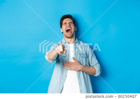 Adult man in casual outfit laughing out loud and pointing at you, looking at something funny, chuckling while standing against blue background 80250487