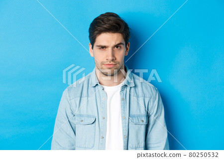 Close-up of suspicious and serious guy raising one eyebrow, looking at camera, standing against blue background 80250532