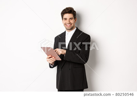 Image of handsome businessman in black suit, using digital tablet and smiling, standing against white background 80250546