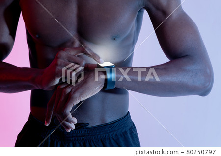 Unrecognizable young black guy with muscular naked torso checking his fitness tracker in neon light 80250797