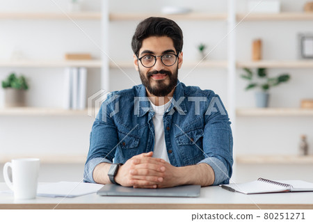 Portrait of confident arab self-entrepreneur man sitting at desk in home office and smiling to camera, copy space 80251271