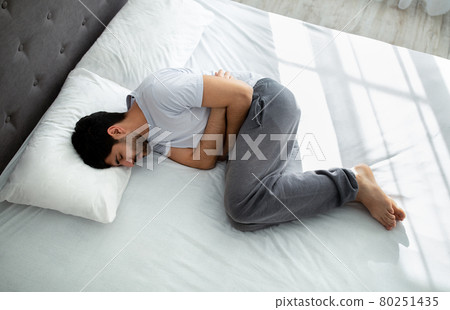 Stomachache. Young arab guy suffering abdominal pain touching aching stomach while lying in bed, above view Stomachache. Young arab guy suffering abdominal pain touching aching stomach while lying in bed, above view 80251435