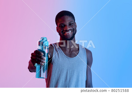 Smiling young black sportsman showing bottle with fresh water in neon light. Fitness, active lifestyle, wellness concept Smiling young black sportsman showing bottle with fresh water in neon light. Fitness, active lifestyle, wellness concept 80251588